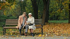 Elderly couple sitting reading a letter together on a bench in an autumn park then smiling and hugging each other