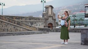 A young woman with a baby in a sling travels to Europe. Standing on the square near the ocean takes on the phone attractions and is online broadcast for your blog