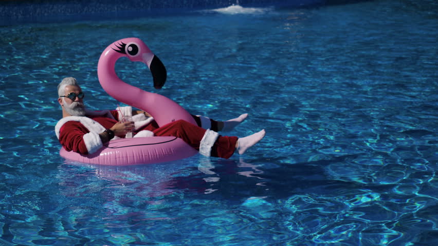 Handsome Santa floating in flamingo inflatable ring into the swimming pool with cocktail glass in hand, chilling and enjoying tropical New Year. Pleasure and happiness concept. Inspiration background Royalty-Free Stock Footage #1018481269