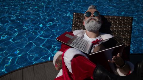 Merry Christmas comic concept in the summertime country. Funny Santa sunbathing on the bad near with swimming pool and use laptop like a reflection. Background of offline vacation without technology