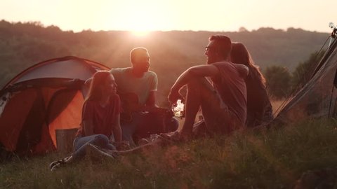 Group of young people camping, talking to each other and listening to a man playing the guitar outside tents. Friendship concept, feeling good, hiking. Nature, excitement