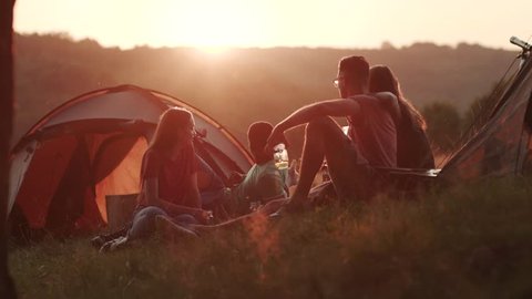 Four friends having rest by their tourist tents in bright sunset light. Mountains, hiking, having fun. Leisure time, friendship concept, tourism. Excitement, relaxing time