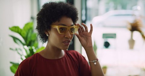Chic black woman looking at row of sunglasses and trying on yellow sunglasses in interior boutique clothing store with soft day lighting. Close up shot on 4k RED camera.