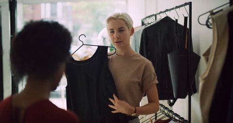 Two female friends perusing clothing on hangers and holding dresses up to each other in interior boutique clothing store with soft day lighting. Close up to Medium shot on 4k RED camera.