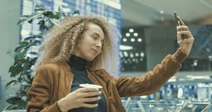 Young woman making selfie by smartphone in airport or station. Portrait of curly girl drinking coffee and having fun in departure terminal. 4k video shot with hands