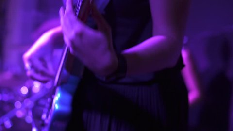 A blonde girl performer playing bass guitar and dancing on the stage under purple light