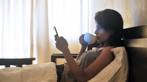 A young woman checking her mobile  phone and taking sip of a coffee while lying on a bed in the morning. A beautiful girl using phone to check email after waking up in the morning in the bedroom