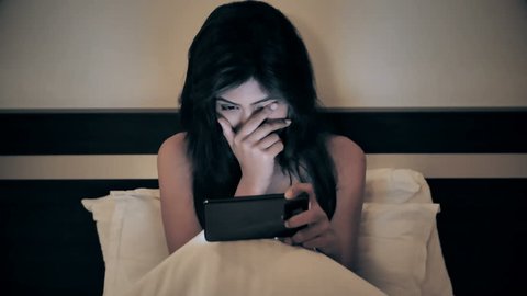 A young and beautiful woman sitting on bed and watching an action or thriller movie on a mobile phone. An attractive girl covering her face while watching a horror film on smartphone or cellphone.