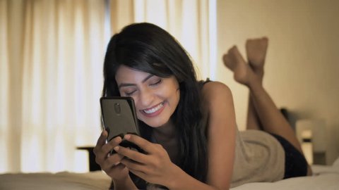 A happy and beautiful woman lying on a bed and talking on video call by using a mobile phone. An attractive girl having a video chat with a boyfriend on a smartphone or cellphone with a smile 
