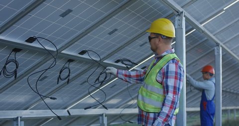 Medium shot of two electrical workers in hardhats connecting wires of solar panels at a solar farm