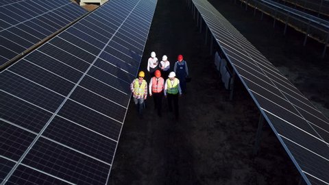 Aerial zoom shot of six electrical workers walking in between long rows of photovoltaic solar panels