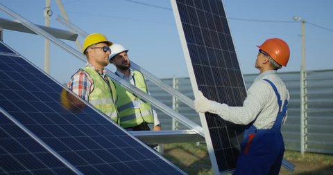 Medium shot of three workers in a uniform and hardhat installing photovoltaic panels on a metal basis on a solar farm