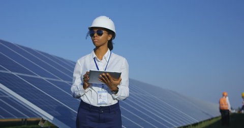 Medium shot of a female electrical worker typing on her tablet inbetween long rows of photovoltaic solar panels