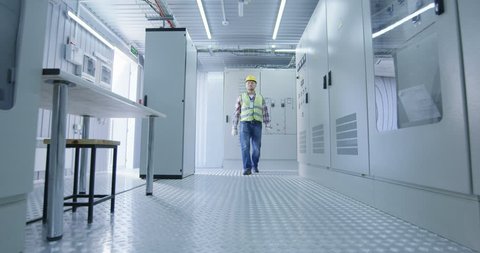 Low angle shot of a electrical worker walking down the hall wearing a hardhat and reflective vest in the control room of an electrical station
