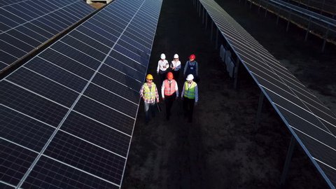 Aerial zoom shot of six electrical workers walking in between long rows of photovoltaic solar panels