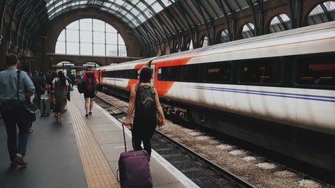 Girl walking through the platform at the station carrying a suitcase and wearing headphones around other people, also walking at the train station. : vidéo de stock