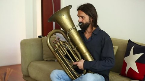 Stock footage of joyful Caucasian man with long hair and beard playing old weary tin tuba in home interior, sitting on sofa, making funny faces.