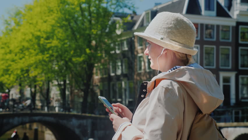 A woman with a smartphone in her hands and headphones is standing near a beautiful canal in Amsterdam on a clear spring day