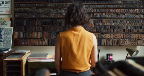 Female artisan typesetter looking at a wall of movable type letters in creative studio with soft day lighting. Medium shot on 4k RED camera.