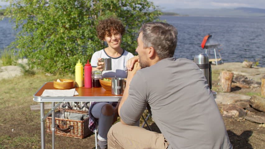 Caucasian couple sitting at picnic table, drinking tea or water from thermo cups and having relaxed conversation by peaceful lake