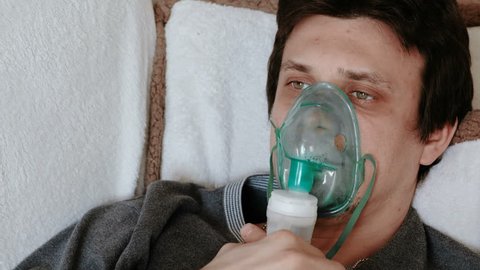 Use nebulizer and inhaler for the treatment. Closeup man's face inhaling through inhaler mask lying on the couch. Front view