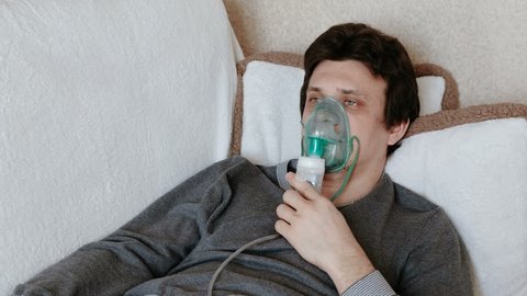 Use nebulizer and inhaler for the treatment. Young man inhaling through inhaler mask lying on the couch. Front view