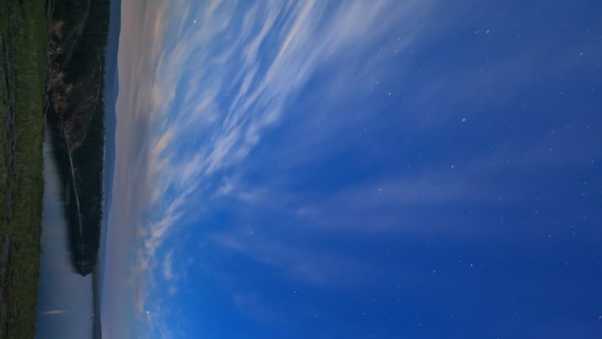 Clouds and stars on a moonlit night. A time-lapse view of a starry sky over a calm body of water and a dark rocky shoreline. The night sky is filled with bright stars that transition into long, fading star trails, creating sweeping lines of light across the deep blue canvas. Wisps of clouds drift slowly through the frame, illuminated by the approaching moonlight. A rocky landmass with patches of green grass sits on the left side of the composition, providing a dark silhouette against the glowing sky. The camera remains completely static throughout the sequence, capturing the natural motion of the celestial bodies and the shifting glow. As the clip progresses, a bright light source, the moon, appears on the horizon, casting an intense glow and creating a subtle lens flare that washes over the scene. The color palette shifts from a rich, dark blue to a pale, luminous blue-white as the moonlight intensifies. The scene captures a serene and majestic night in a remote natural setting, highlighting the beauty of the cosmos and the stillness of the landscape. The gradual accumulation of light creates a dynamic visual narrative, moving from the quiet darkness of early night to the brilliant illumination of moonrise. The long-exposure technique reveals the earth