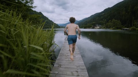 Young millennial authentic hipster man with body tattoos runs fast on wooden boardwalk in amazing natural surroundings, mountain alpine lake with clean fresh word. jumps in funny bomb way