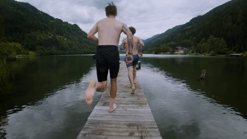 Two friends on summer vacation or holiday run on wooden boardwalk on alpine mountain lake, jump into cold fresh clean water to get refreshed in heat, natural outdoor lifestyle