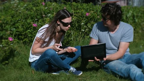 Young couple man and woman are working together on mobile and laptop sitting in park.