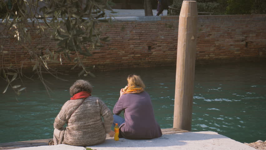 two women having a snack on a canal in Venice, sitting on the quay