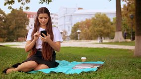 young woman has online video chat with friend waving hand for greeting. cheerful girl using smartphone sitting on lawn in park