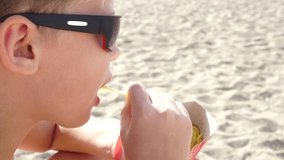Closeup profile portrait of cute white little kid eating potato chips with appetite as snack on sandy summer beach outside. Real time 4k video footage.