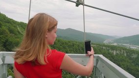 Young blonde in red T-shirt broadcasts video through smartphone with broken screen on bridge against background of green mountain valley and two people on zipline attraction - Powered by Shutterstock - Get 15% off with code: PIKWIZARD15
