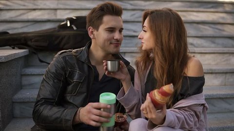 Young couple sitting on the stairs and eating croissants, drinking coffee outdoors, sitting on the plaid and talking. Loving couple spending time together and laughing