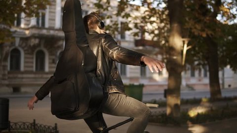 Brunette handsome young man in black casual clothes, headphones riding a bike by city park with guitar in black case on the back and listening to the musicwith outstretched hands. Green trees around