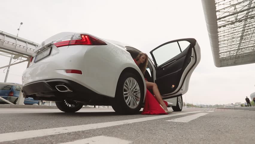 Fashion Model In Red Dress And High Heel Shoes Is Goes Out Of The White Car. Airport Terminal.