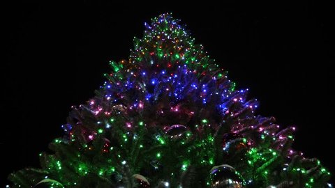 shining high Christmas fir-tree decorated with garlands and multi-colored balls against night sky. little snow falls