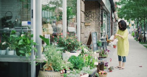 Female store worker going outside boutique floral shop to water plants in exterior with soft day lighting. Medium shot on 4k RED camera.