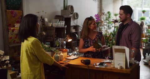 Woman purchases orchid flower with boyfriend using credit card tap in interior boutique plant shop with soft day lighting. Medium shot on 4k RED camera.