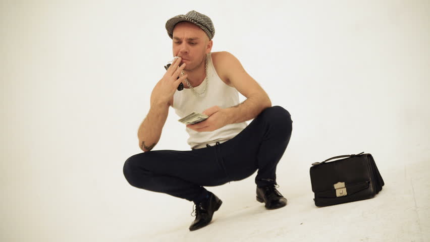 An old fashioned squatting slav in a tank top and a baker boy cap sitting with a cigarette throwing money away Royalty-Free Stock Footage #1018635466
