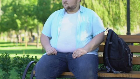Funny chubby male sitting on bench in park and curiously looking at passersby