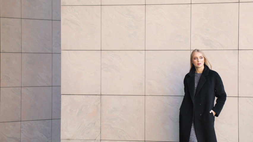 Attractive caucasian girl walking confidently before wall, wearing black coat and gray dress on warm day