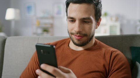 Happy Young Man Uses Smartphone while Sitting on a Sofa at Home. Man Browses Through Internet, Watches Videos and Uses Social Networks at Home.