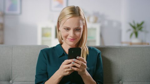 Beautiful Blonde Woman Uses Mobile Phone while Sitting on a Sofa at Home. Smiling Happy Woman Uses Smartphone for Browsing through Internet, Social Networks and Watching Videos.