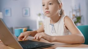 Smart Little Girl Does Homework in Her Living Room. She's Sitting at Her Desk, Uses Laptop and Writes with a Pen in Her Textbooks.