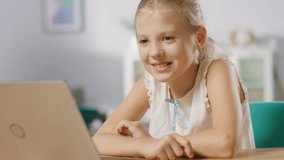Cute Little Girl Uses Laptop While Sitting at the Desk in the Living Room. Child Has Fun while Browses Through Internet and Watches Cartoons and Funny Videos.