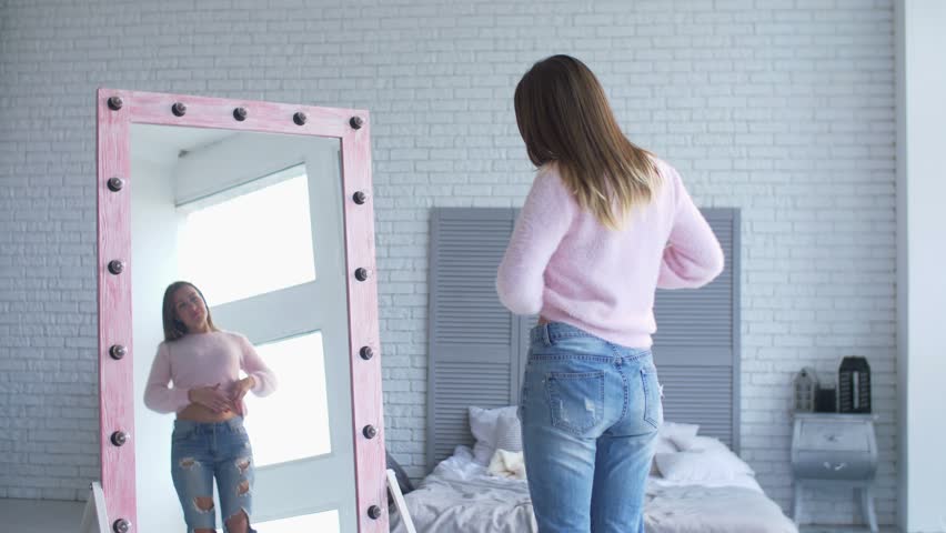 Beautiful adult woman in loose panrs admiring her body shape and appearance after losing weight while standing in front of big mirror in domestic room. Happy female showing weight loss success. Royalty-Free Stock Footage #1018671055