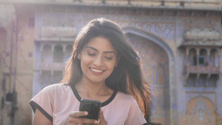 A beautiful smiling girl walking on a city street and using mobile phone while flock of pigeons flying in the background. A moving and back lit shot of an attractive woman on a smartphone smiles Royalty-Free Stock Footage #1018672180