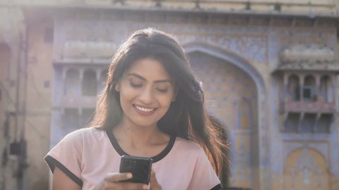 A beautiful smiling girl walking on a city street and using mobile phone while flock of pigeons flying in the background. A moving and back lit shot of an attractive woman on a smartphone smiles